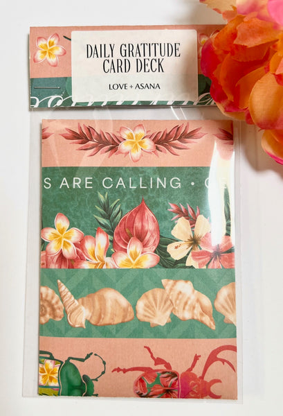 A packaged card deck labeled 'DAILY GRATITUDE CARD DECK' by Love & Asana, featuring tropical floral and shell illustrations on peach and green panels, photographed on a white surface with a coral flower prop at the top right.