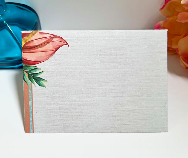 A square white textured card or notepad with a watercolor floral design in the top-left corner, featuring a coral-pink leaf, green leaves, and a peach stripe, photographed on a white surface with decorative props.