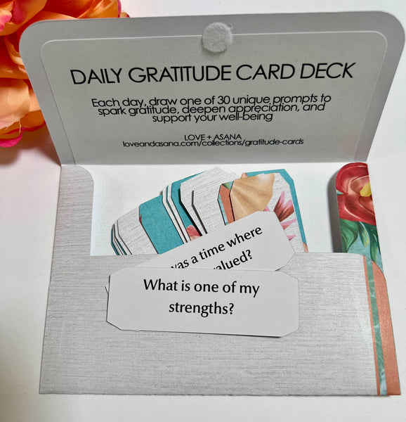 An open daily gratitude card deck in a silver envelope, showing sample cards with prompts like 'What is one of my strengths?' and colorful illustrated card edges. The card deck packaging at the top reads 'DAILY GRATITUDE CARD DECK' with a website URL.