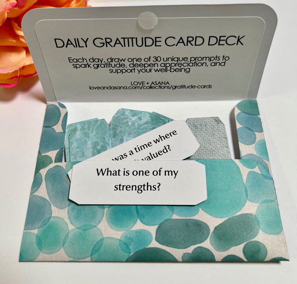 An open Daily Gratitude Card Deck in a teal and white patterned envelope, showing several sample gratitude cards inside, with the back cover visible in the background.
