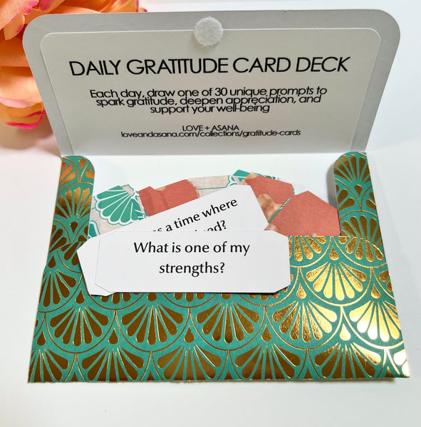 An open daily gratitude card deck with a white header card reading 'DAILY GRATITUDE CARD DECK' and teal-and-gold patterned cardstock inside. A sample prompt card is visible in the center.